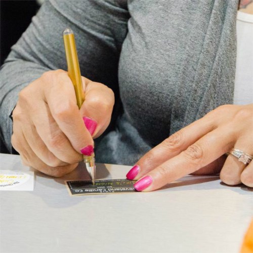 A person with pink nails writes on a small card with a pen, resting their hand on a table.