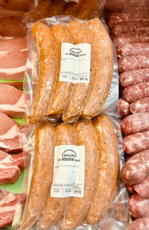 Two packages of boudin sausage in clear plastic, displayed on a butcher's counter with other meats in the background.