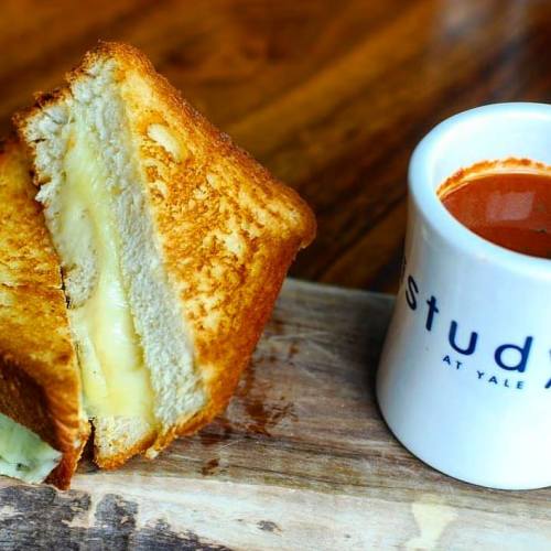 A golden grilled cheese sandwich with melted cheese, served alongside a cup of tomato soup on a wooden surface.