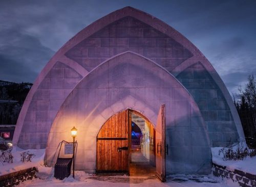 A large, arching ice structure with a wooden door, illuminated by a lamp, set against a twilight sky. Snow covers the ground.