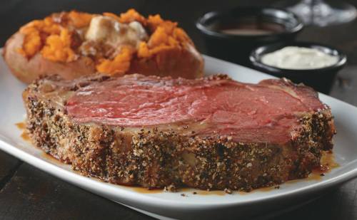 A plate featuring a juicy prime rib steak with a baked sweet potato topped with butter and two dipping sauces.