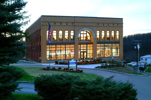 A brick building with large windows, surrounded by greenery and flowers, illuminated at dusk. An American flag is visible.