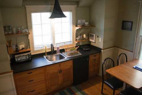 A cozy kitchen with wooden cabinets, a sink, a toaster, and a dining table with black chairs near a window.