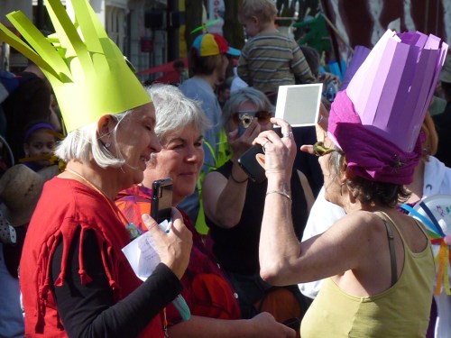 Three women wearing colorful paper hats engage in conversation at a lively outdoor event, with a crowd in the background.