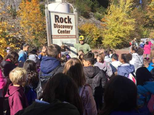 A group of children gathers around a sign for the Rock Discovery Center, surrounded by autumn foliage.