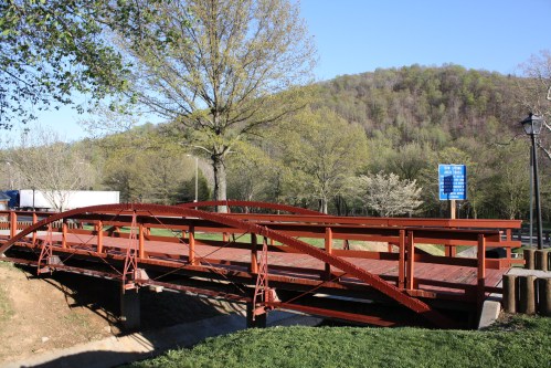 A red metal bridge spans a small creek, surrounded by trees and a hillside under a clear blue sky.