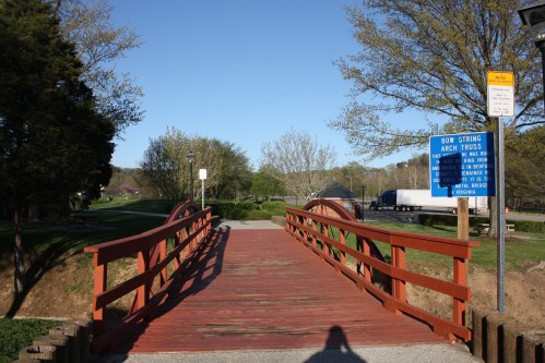 A wooden bridge with a red railing spans a path in a park, surrounded by trees and a clear blue sky.