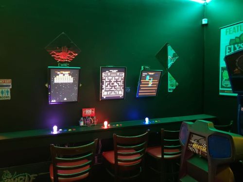 A retro arcade setup with three screens displaying classic games, colorful lights, and empty chairs in a dark room.