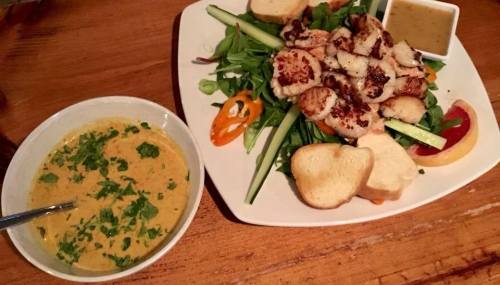 A plate of grilled chicken on a bed of greens with vegetables, accompanied by a bowl of creamy soup and bread slices.