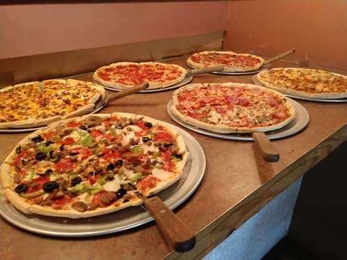 A variety of pizzas on metal trays, including pepperoni, veggie, and meat lovers, displayed on a countertop.