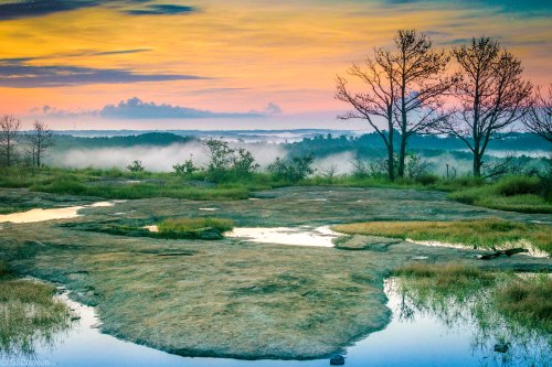 A serene landscape at sunrise, featuring misty hills, a rocky foreground, and silhouetted trees against a colorful sky.