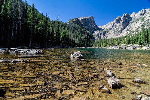 A serene lake surrounded by tall pine trees and rocky mountains under a clear blue sky.