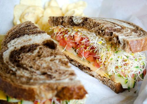 A close-up of a sliced sandwich with sprouts, tomatoes, and cheese on dark rye bread, served with potato chips.