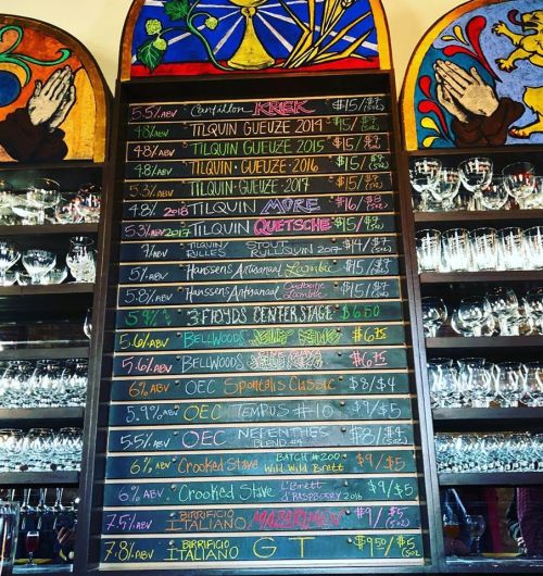 Chalkboard menu displaying various beers and prices, with colorful stained glass in the background.
