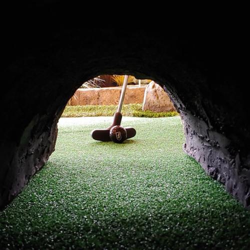 A view through a cave-like opening, revealing a putting green and a golf club resting on the artificial grass.