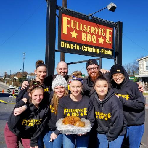 A group of smiling staff members in hoodies pose in front of a BBQ restaurant sign, holding a platter of food.