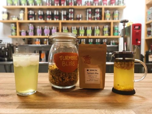 A glass of iced tea, a jar of turmeric bliss tea, and a French press on a wooden table with a colorful background.