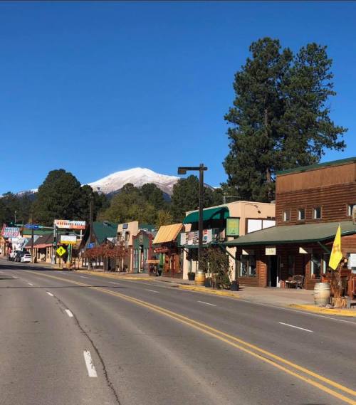 A quiet street lined with shops, with snow-capped mountains in the background under a clear blue sky.