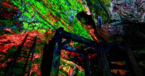 Colorful lights illuminate a rocky cave interior, highlighting wooden structures and a ladder against vibrant stone walls.