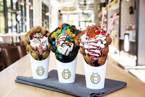Three colorful ice cream desserts in cups, topped with sprinkles, chocolate, and candies, displayed on a table.