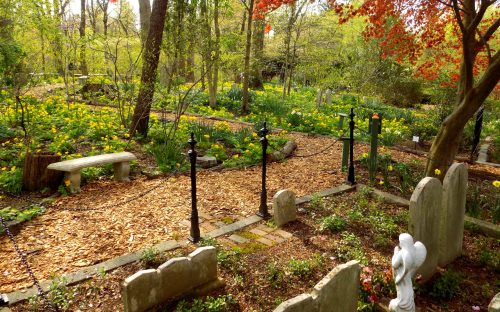 A serene garden with a path, surrounded by flowers, gravestones, and a statue of an angel under a bright sky.