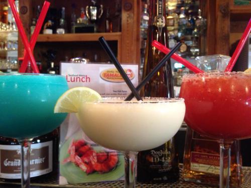 Three colorful cocktails with straws, garnished with lime, in front of a menu and bottles on a bar counter.