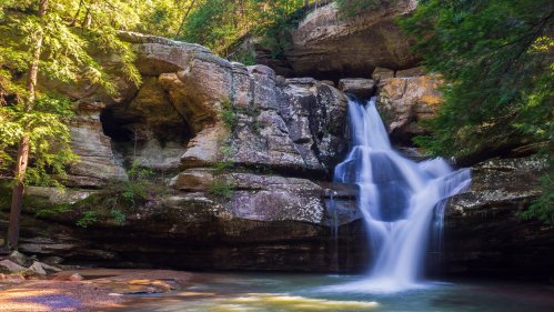 A serene waterfall cascades over rocky cliffs, surrounded by lush green trees in a tranquil forest setting.