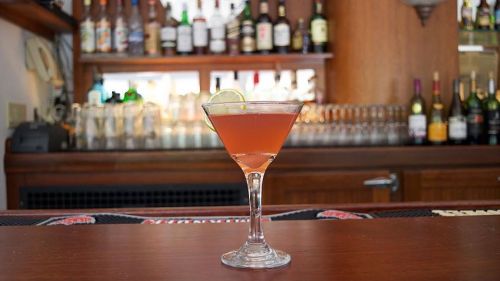 A cocktail in a martini glass garnished with a lime slice, set against a bar with various bottles in the background.