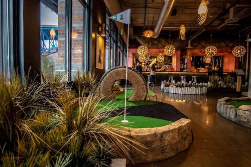 Indoor mini-golf course featuring a stone archway, vibrant greenery, and a bar in the background with unique lighting.