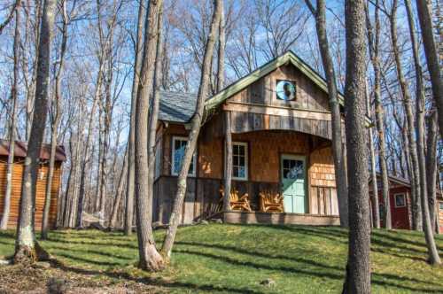 A cozy wooden cabin nestled among trees, featuring a green roof and a light blue door, with two chairs on the porch.