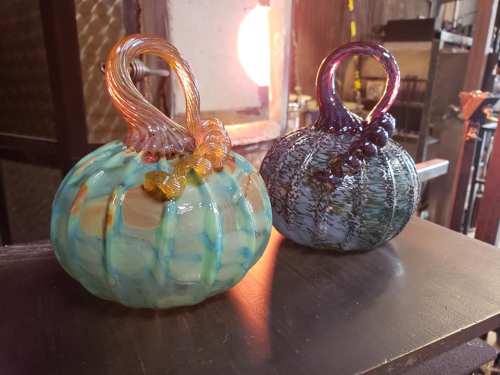 Two colorful glass pumpkins with unique textures and handles, displayed in a workshop with a glowing furnace in the background.