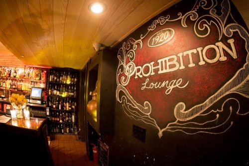 A cozy bar interior featuring a chalkboard mural that reads "Prohibition Lounge" with shelves of liquor in the background.