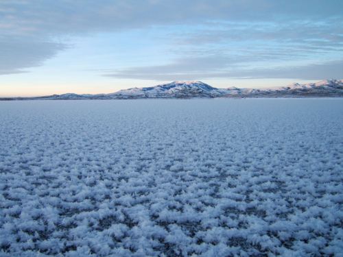 A vast, snowy landscape with a flat, white surface and distant mountains under a soft blue sky.