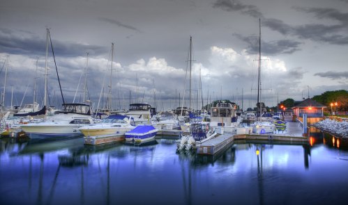 A serene marina at dusk, featuring various boats and yachts reflected in calm water under a cloudy sky.