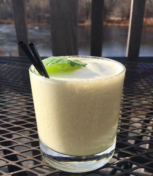 A creamy cocktail in a glass with two straws and a mint leaf, set on a table by a river.