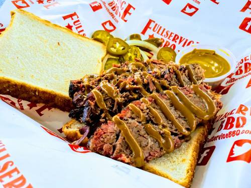 A plate featuring sliced smoked brisket on white bread, topped with mustard and served with pickles and jalapeños.
