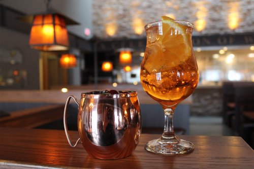 Two cocktails on a wooden bar: a copper mug with berries and a tall glass with orange slices and bubbles. Warm lighting in background.