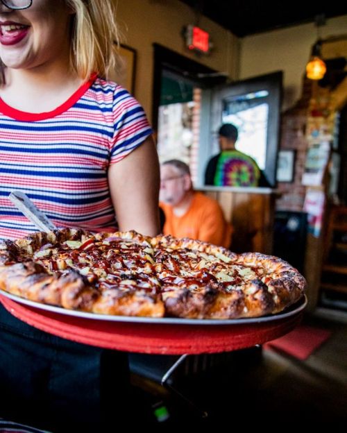 A server holds a BBQ pizza with pineapple, while a diner sits in the background of a cozy restaurant.