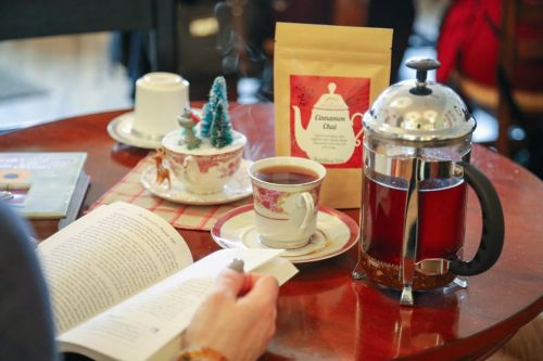 A cozy scene with a cup of tea, a French press, a book, and a packet of cinnamon chai on a wooden table.