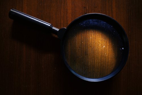 A magnifying glass resting on a wooden surface, casting a circular light and revealing dust particles underneath.
