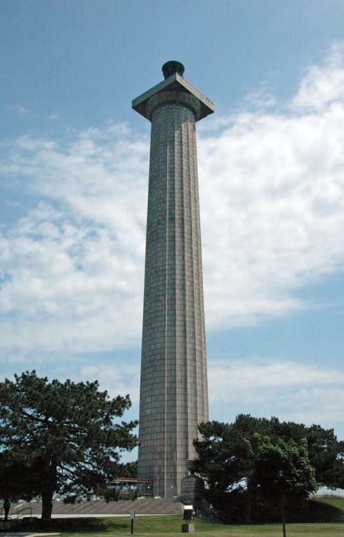 A tall stone monument with a square base, surrounded by trees under a partly cloudy sky.