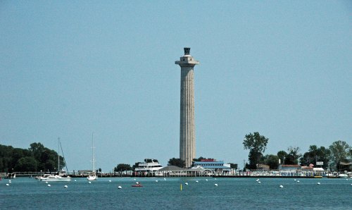 A tall stone tower stands by the water, surrounded by trees and boats on a clear day.