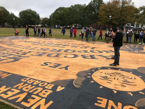 A large wooden mural on the ground with text and a sun design, surrounded by people in a park on a cloudy day.