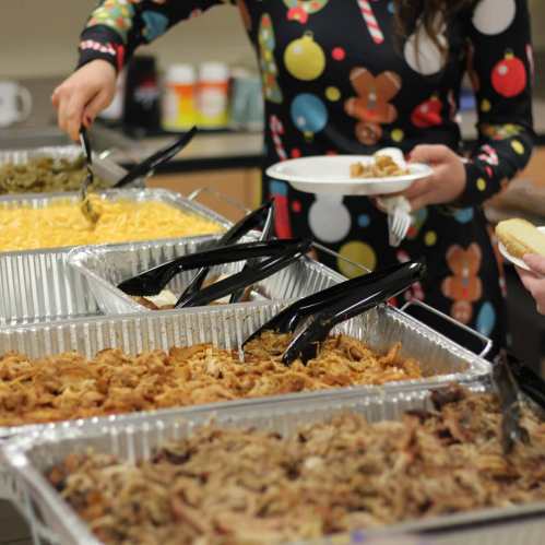 A person serves food from a buffet with various dishes, including macaroni and pulled pork, while holding a plate.