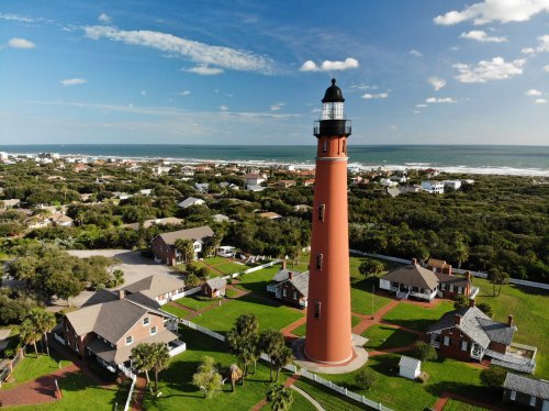 Aerial view of a tall red lighthouse surrounded by green lawns and houses near the ocean under a blue sky.