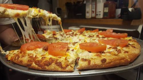 A close-up of a cheesy pizza with tomato slices, being pulled from a tray, showcasing its gooey cheese and golden crust.