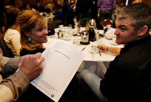 A woman and a man sit at a dining table, with a hand holding a clipboard in the foreground, surrounded by other diners.