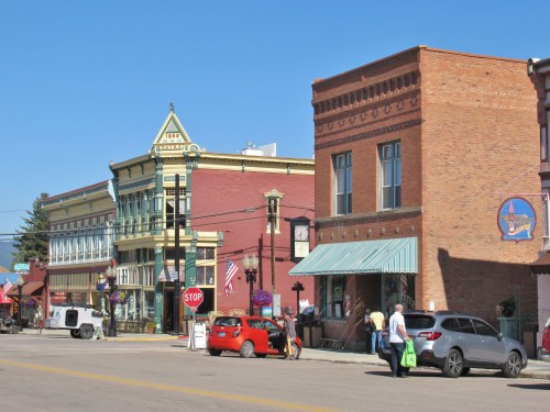 Historic buildings line a sunny street, featuring colorful facades, flags, and people walking by.