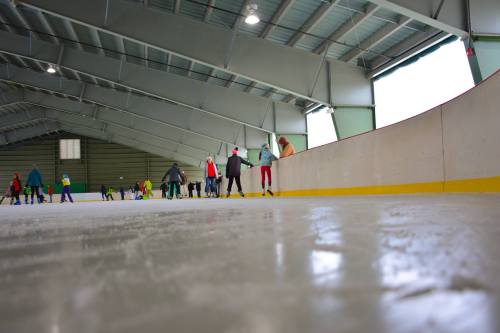 A view of an indoor ice skating rink with people skating and holding onto the edge for support.