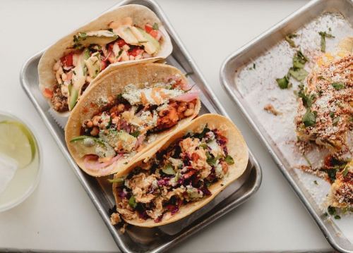 Three delicious tacos on a metal tray, filled with various toppings and garnished with cilantro, alongside a drink.